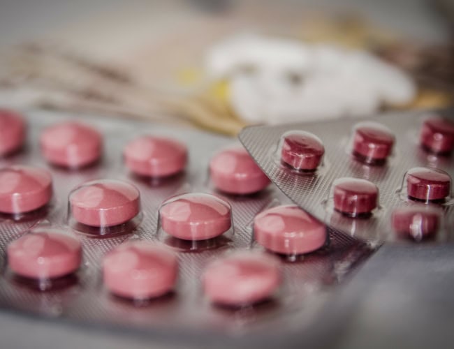 Close-up of several blister packs containing pink round tablets. The packs overlap slightly, showing some tablets in foil and others exposed. The background is blurred, containing off-focus objects that hint at a medical or pharmaceutical setting.