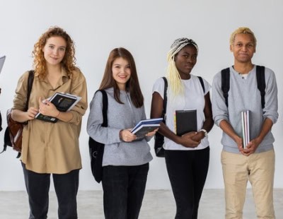 A group of six people are standing against a plain wall, smiling. They are casually dressed and holding books, notebooks, and a tablet. The diverse group appears to be students or colleagues, each with a unique style and demeanor.