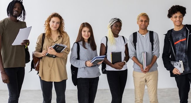 A group of six people are standing against a plain wall, smiling. They are casually dressed and holding books, notebooks, and a tablet. The diverse group appears to be students or colleagues, each with a unique style and demeanor.