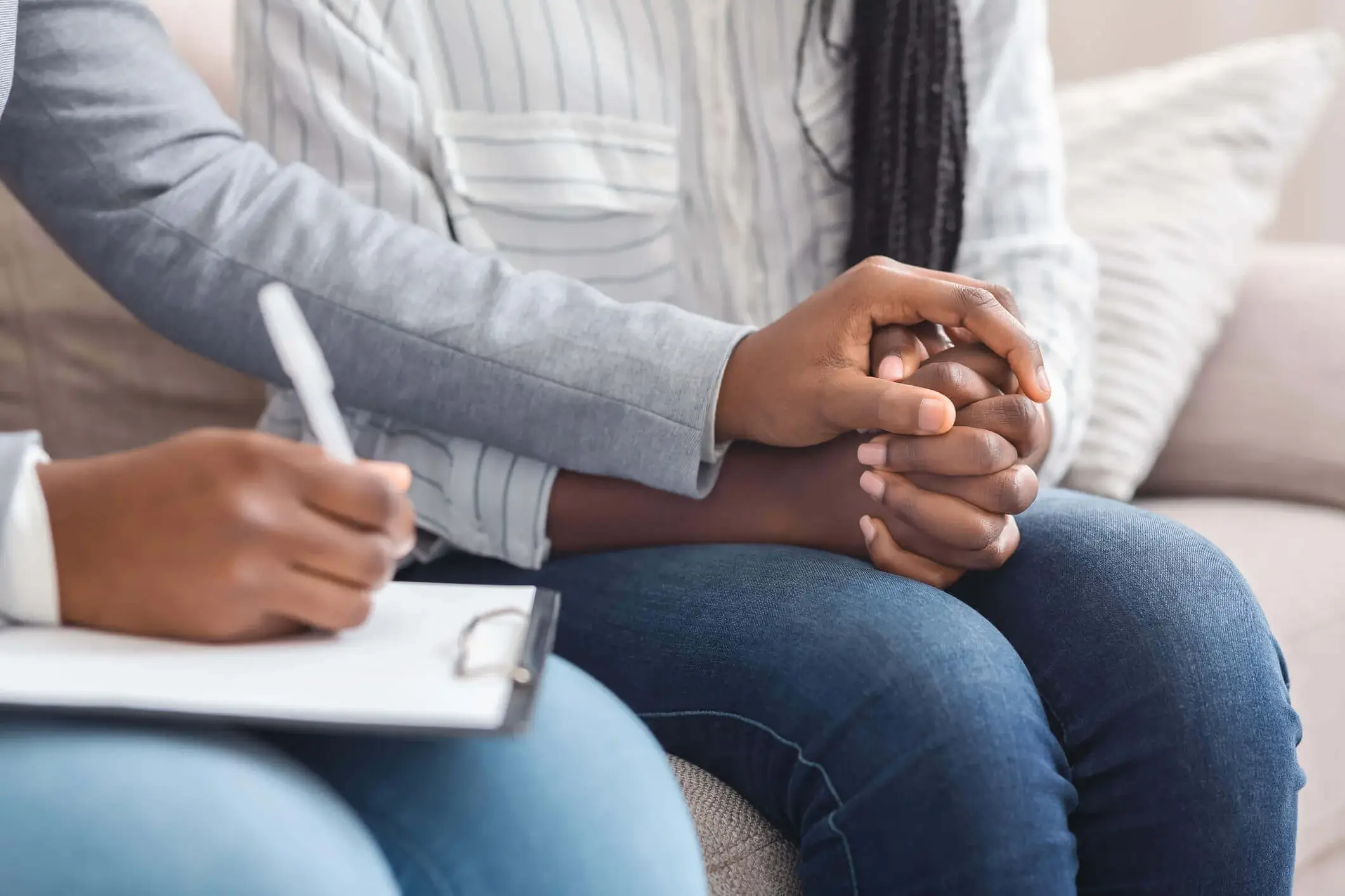 Two people are sitting closely on a couch. One person is holding the other's hand in a comforting gesture. The person next to them is writing on a clipboard, suggesting a supportive or counseling environment. Both individuals are wearing casual clothing.