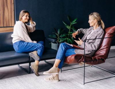 Two women engaged in a conversation are seated in a modern room with stylish furniture and a potted plant in the background. One woman is sitting on a black sofa, smiling and touching her hair, while the other sits on a brown leather chair, gesturing with her hands.