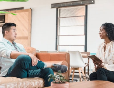 A man and a woman are sitting in a modern office space, engaged in a conversation. The man is sitting on a tan leather couch, while the woman holds a tablet, possibly conducting a depression screening near me. There are plants and a large window in the background.