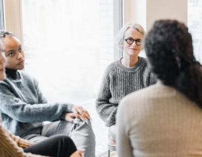 A diverse group of four people sit together in a circle, engaged in conversation. They are indoors, with soft natural light coming through the windows. The atmosphere seems relaxed and friendly. One person, an older woman with glasses, appears to be speaking.