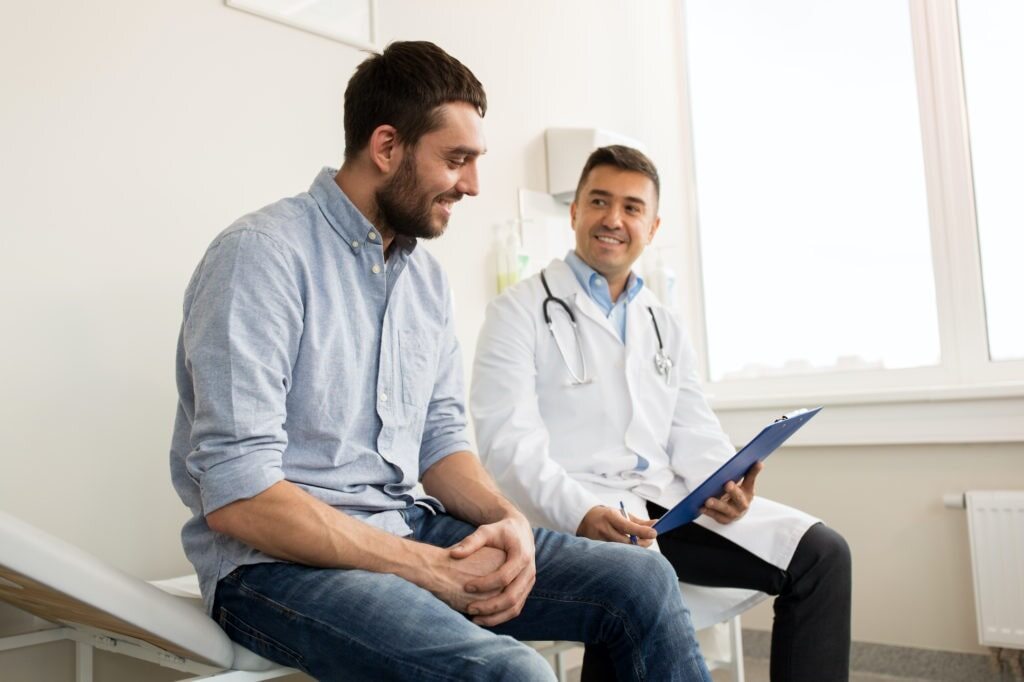 A man sits on an examination table, smiling and conversing with a doctor who is seated nearby, holding a clipboard. The doctor, wearing a white coat and a stethoscope around his neck, is also smiling. They are in a bright, modern medical office.