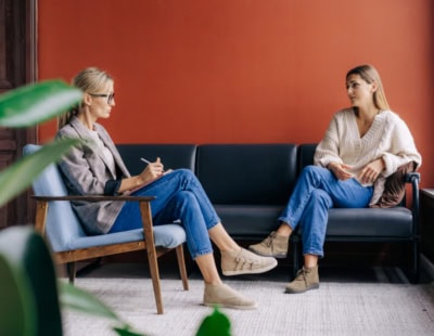 Two women are sitting across from each other in a cozy room. One, in glasses and a blazer, holds a pen and listens attentively, while the other, in a knitted sweater, gestures as she speaks. Both are seated on blue chairs, and there is a potted plant in the foreground.