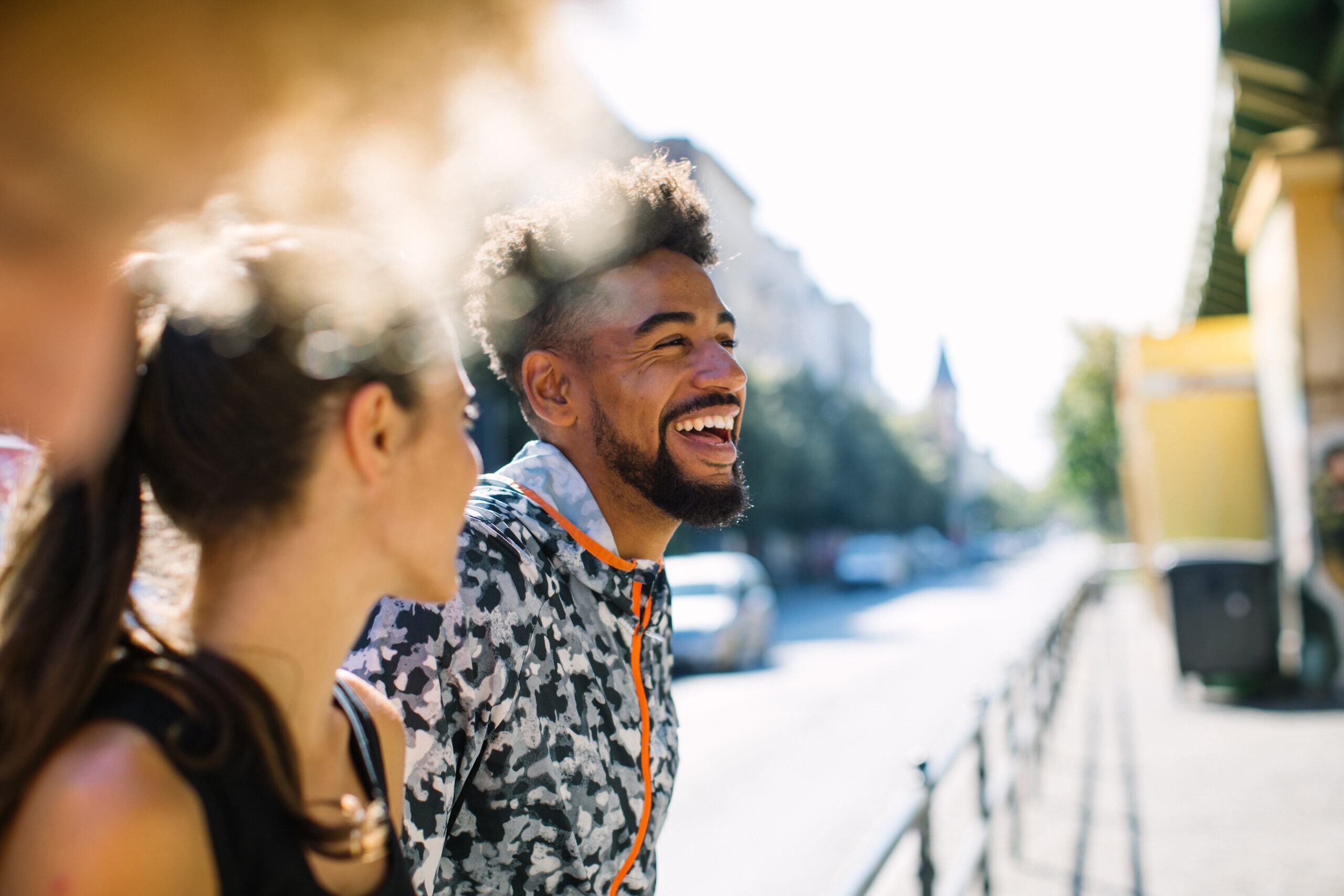 A man and a woman share a laugh while walking down a sunny street. The man wears a patterned jacket and has a beard and curly hair. The woman, with her hair tied back, wears a tank top. They appear relaxed and happy in an urban setting with blurred background.