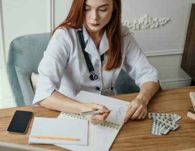 A young woman with red hair, wearing a white lab coat and a stethoscope, sits at a wooden desk writing in a notebook. A smartphone, a pencil, a notepad, and a tray with medical pill packets are also on the desk. An ornate white wall is in the background.