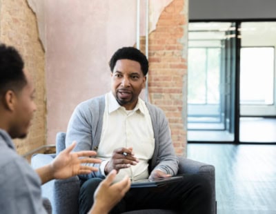 A man sits on a gray couch holding a clipboard and pen, engaging in conversation with another person who is making hand gestures. They are in a room with brick walls and large windows with natural light. The atmosphere appears professional and focused, exploring options for when therapy doesn’t work.