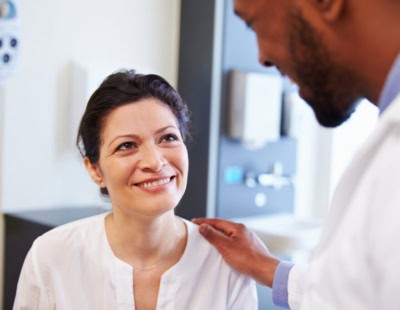 A woman is sitting and smiling up at a man in a white coat, who appears to be a doctor. The doctor, who diagnoses bipolar disorder in adults, has his hand on the woman's shoulder, offering comfort. They are in a medical office setting.