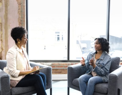 Two women are sitting in gray chairs facing each other in a modern, well-lit room. One woman, holding papers, appears to be listening attentively, while the other woman speaks, gesturing with her hands. Large windows behind them let in ample natural light.