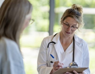 A female doctor with a stethoscope around her neck is writing on a clipboard while speaking to a patient. The doctor wears glasses and a white coat, and the background is softly blurred with greenery visible through a window.