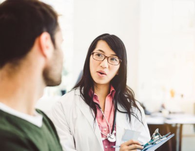 A female doctor in a white coat and pink shirt is holding a clipboard and talking to a male patient with a beard who is wearing a green sweater. They are in a well-lit medical office.