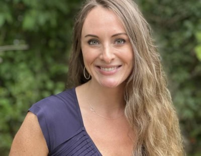 A woman with long wavy hair smiles at the camera, standing outdoors in front of lush green foliage. She is wearing a dark blue dress and hoop earrings, radiating the serene confidence often cultivated through therapy for women in Raleigh.