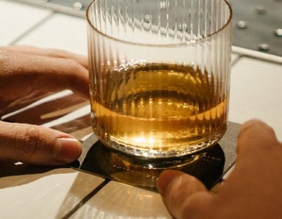 A person places a glass tumbler filled with a golden-brown drink onto a square black coaster on a tiled surface. The tumbler has vertical ridges, and both hands are visible in the image.