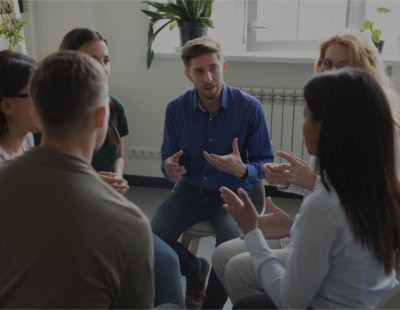 A group of six people is seated in a circle in a well-lit room with large windows and plants on the windowsills. They appear to be engaged in a discussion, with one person in a blue shirt gesturing while others listen and respond.