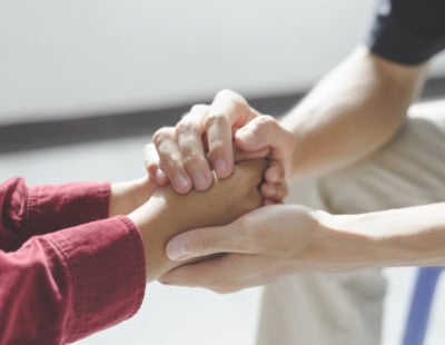 Close-up of two people holding hands. One person, wearing a red long-sleeve shirt, extends their hands as the other person gently grasps them with both hands, providing comfort and support. The background is blurred, emphasizing the connection between the individuals.