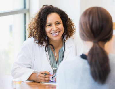 A smiling doctor with curly hair and a white coat sits at a desk, engaging in a conversation with a patient who is facing away from the camera. The doctor has a stethoscope around her neck and appears to be in a well-lit office setting.