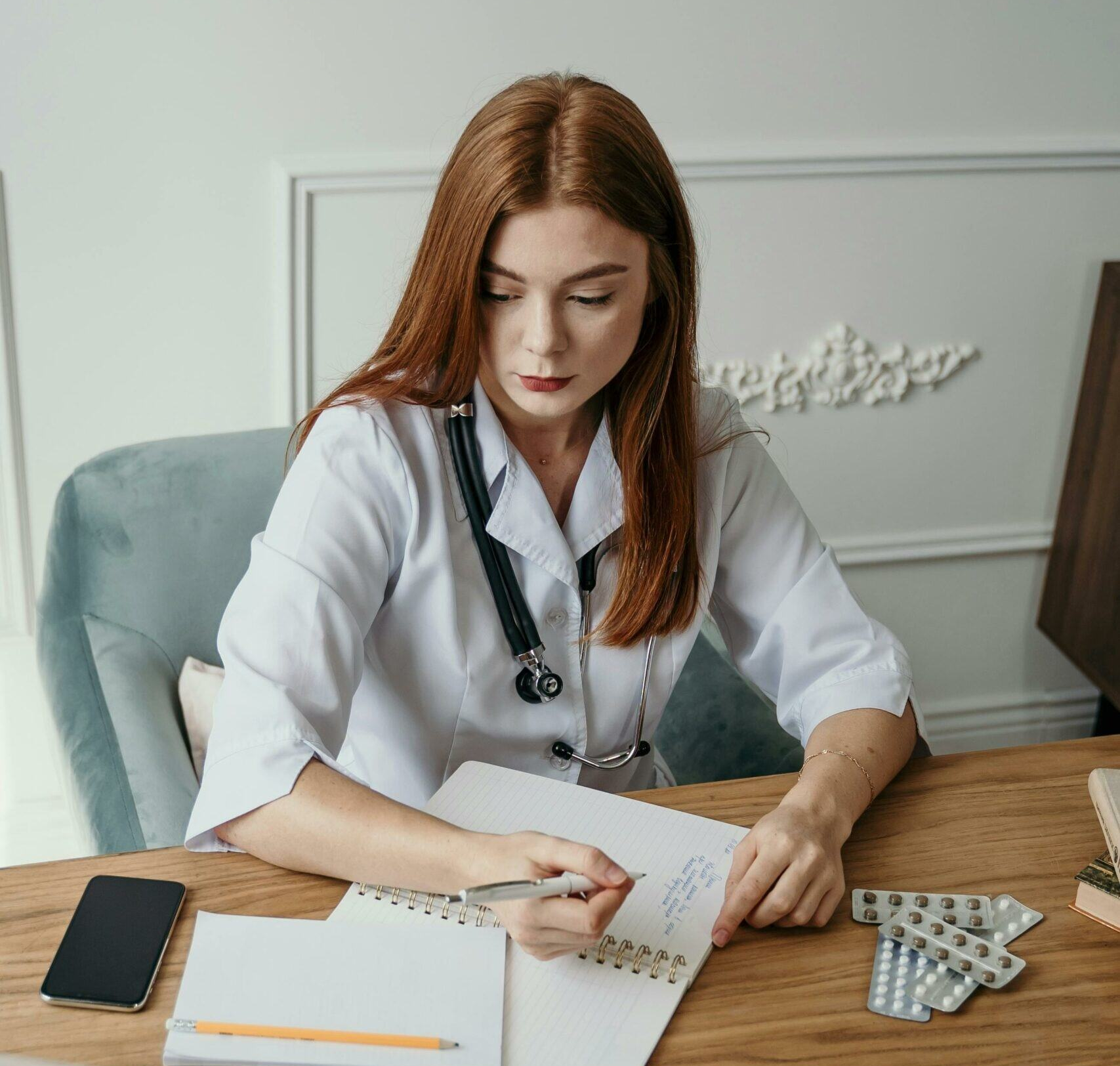 A female doctor with long red hair, wearing a white coat and stethoscope, sits at a wooden desk. She is writing in a notebook with a pen. Nearby are a smartphone, an orange pencil, and several blister packs of medication. She appears focused on her work.