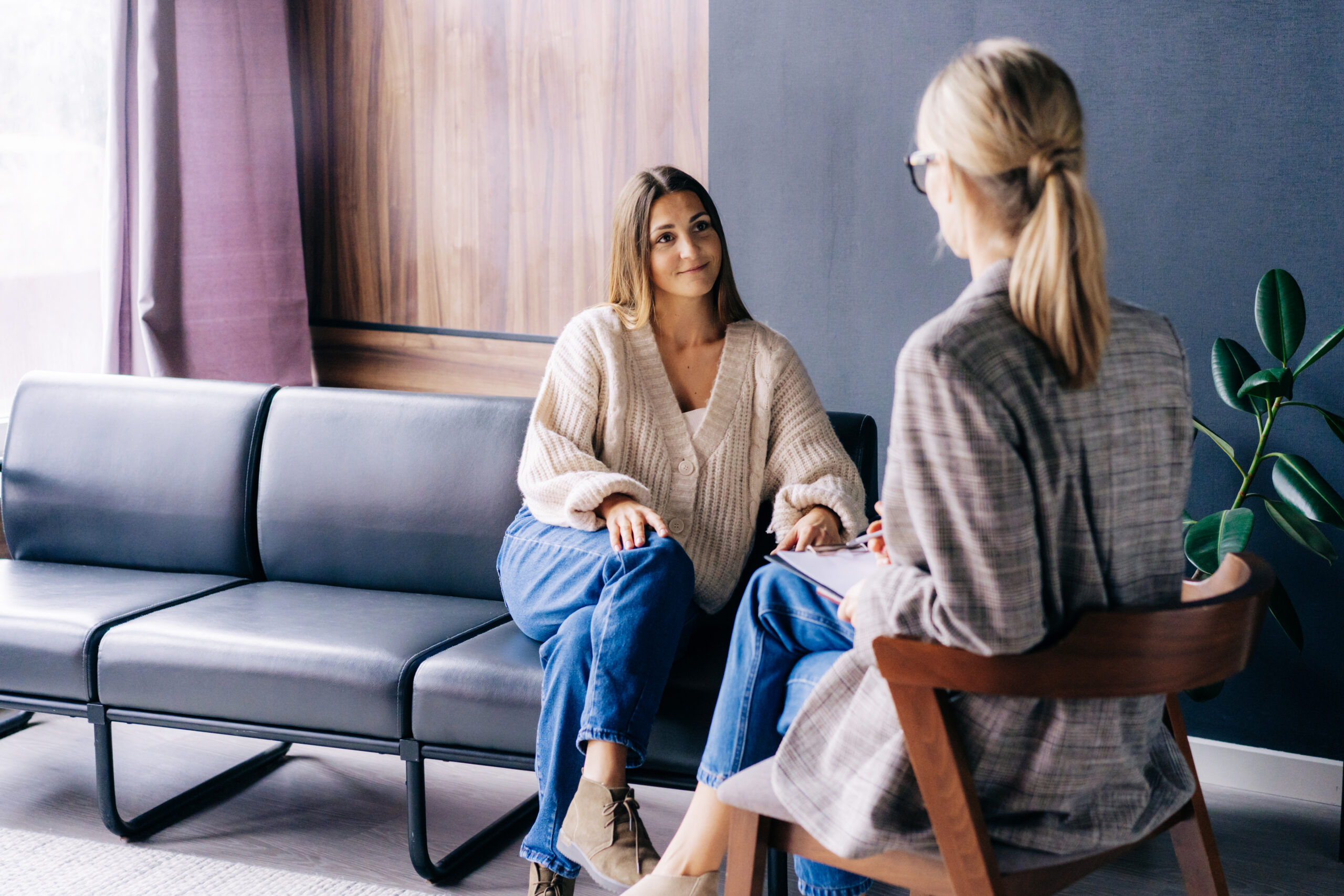 Two women are sitting in a modern, cozy room. One woman with long hair and a light knitted sweater sits on a black leather couch, smiling. The other woman, with blonde hair tied back and wearing glasses and a plaid blazer, sits on a wooden chair, facing her.