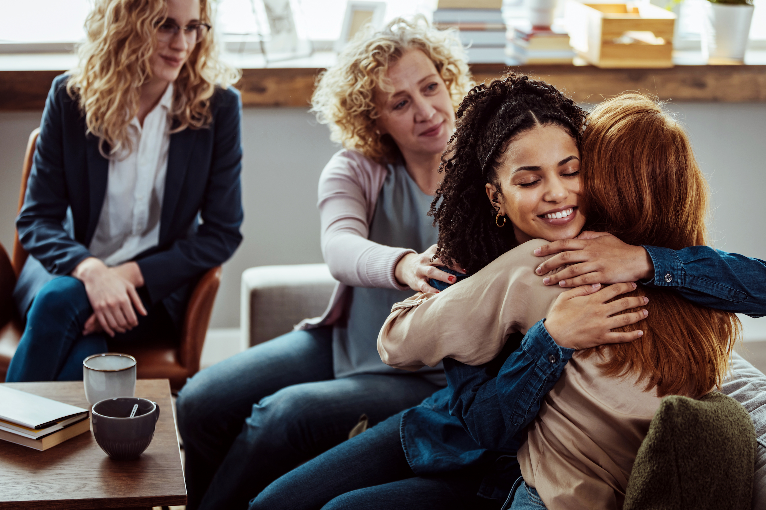 A group of four women is gathered in a cozy room. Two of them are seated on a couch, sharing a warm hug. Another woman sitting nearby reaches out to offer comfort. The fourth woman sits slightly apart, observing the scene with a thoughtful expression. Books and mugs are on the table.