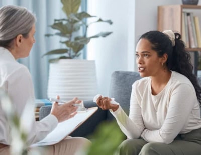 A young woman holds a tissue while sitting on a couch, engaged in conversation with an older woman holding a clipboard. They appear to be in a counseling or therapy session in a well-lit room with a plant and bookshelf in the background.