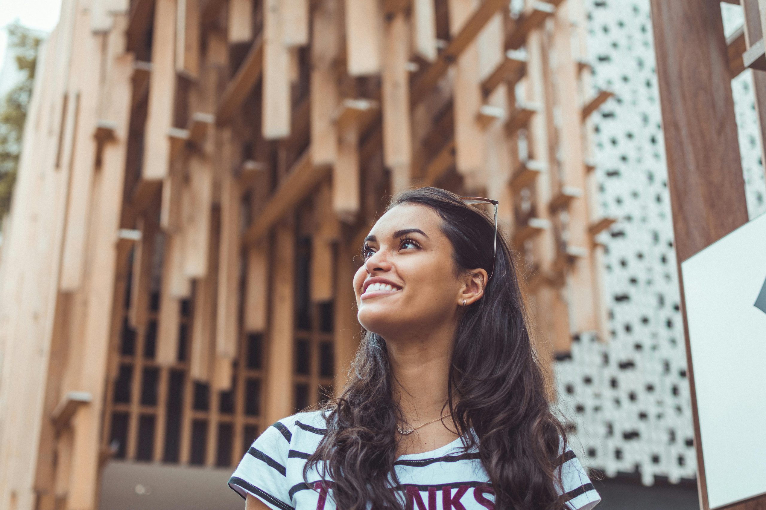 A woman with long dark hair, wearing a striped shirt, looks up and smiles. She stands in front of a modern wooden architectural structure with geometric patterns.