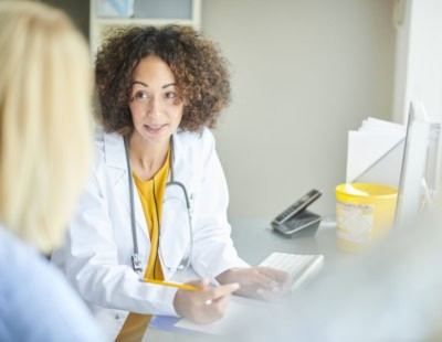 A doctor with curly hair, wearing a white coat and stethoscope, sits at a desk facing a patient in Raleigh. The doctor gestures while discussing treatment for OCD, with a computer keyboard and paperwork on the desk as sunlight filters in from a nearby window.