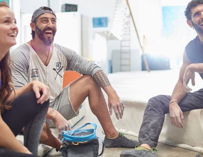 Three people are seated on the floor of an indoor climbing gym, smiling and looking towards the right. They are in casual athletic clothing. Various climbing holds and walls are visible in the background. A blue bag is on the floor in front of them.
