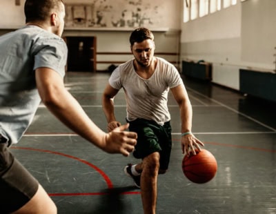 Two men play basketball in an indoor gym. The man in the foreground dribbles the ball with determination, while the other man attempts to defend. The court has red lines and is dimly lit, with windows lining one side of the gym.