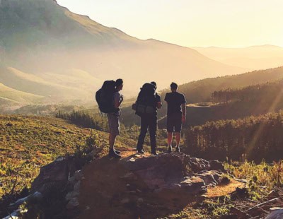Three hikers with backpacks stand on a rocky outcrop, overlooking a vast mountain landscape bathed in the warm light of a setting sun. The rolling hills and distant peaks are partially covered in mist, creating a serene and picturesque scene.