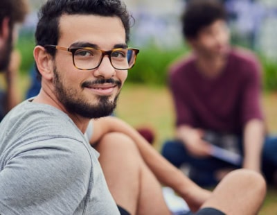 A person with glasses and a beard is sitting outside, smiling and looking at the camera, happy to have managed his anxiety. They are wearing a grey shirt. In the blurred background, there are other people sitting on the grass, engaged in conversation.