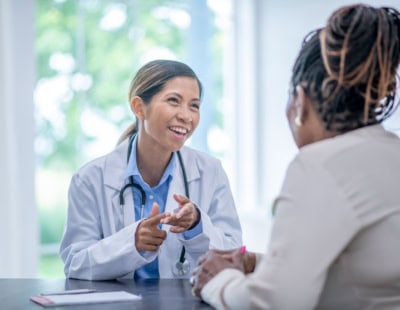 A female doctor in a white coat, smiling and conversing with a seated female patient. The doctor has a stethoscope around her neck and gestures with her hands. The background is bright with green foliage visible through a window.