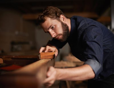 A bearded man is focused intently on his woodworking project, carefully sanding a piece of wood in a dimly lit workshop. He is wearing a denim shirt, and various woodworking tools and equipment are visible in the blurred background.