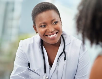 A smiling healthcare professional with short hair and a white coat, and a stethoscope around their neck, listens attentively to a patient. The background is blurred to emphasize the interaction.