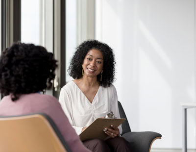 A woman with curly black hair, wearing a light-colored blouse, sits and holds a clipboard while smiling and talking to another person with curly black hair, wearing a pink top. They are seated in modern chairs near large windows.