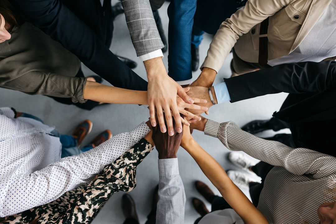 A diverse group of people standing in a circle place their hands together in the center, symbolizing unity and teamwork. The image showcases varied skin tones, clothing styles, and accessories, highlighting inclusivity and collaboration.