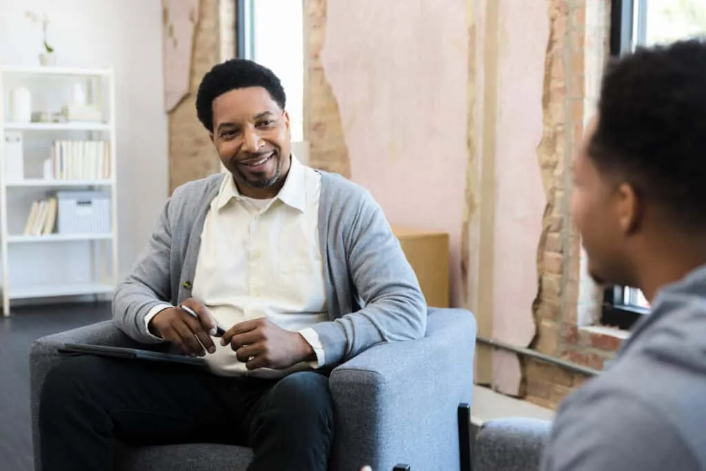 Meeting one-on-one, the mid adult male therapist and his young adult male patient smile at each other.
