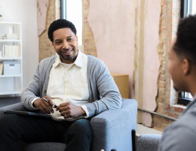 Meeting one-on-one, the mid adult male therapist and his young adult male patient smile at each other.