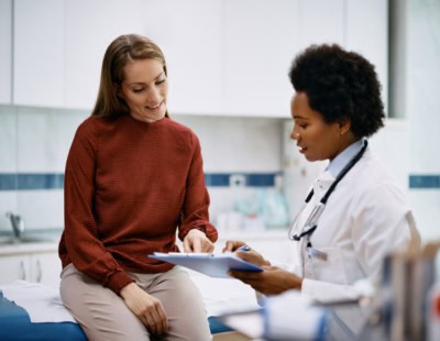 A woman wearing a red blouse is sitting on an examination table in a medical office, smiling, and pointing at a clipboard. A healthcare professional, dressed in a white coat with a stethoscope around their neck, is attentively looking at the clipboard and taking notes.