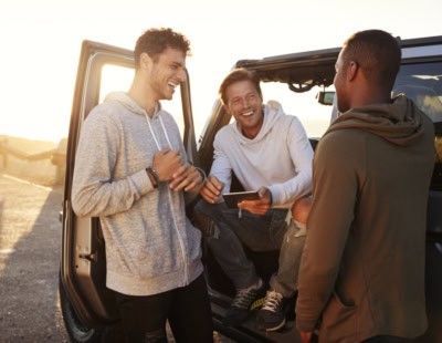 Three friends enjoying a conversation next to an off-road vehicle at sunset. Two men are standing, one in a gray hoodie and the other in a green hoodie, while the third man sits in the vehicle's open door, wearing a white hoodie, and holds a tablet.