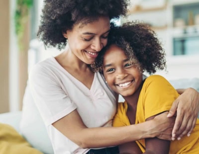 A woman in a white shirt is smiling as she holds and hugs a young female child in a yellow shirt.