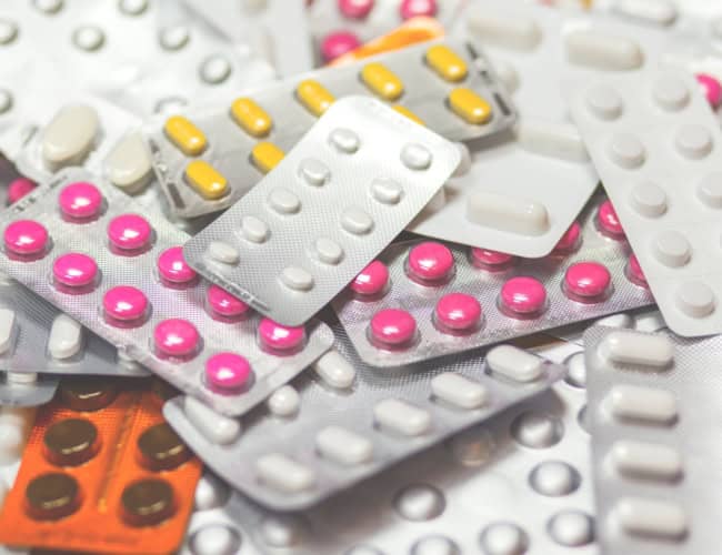 A diverse assortment of medicine blister packs, filled with tablets and capsules of various colors like pink, yellow, white, and brown, is scattered randomly—each potentially a vital component in mental health treatment for college students.
