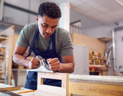 A man wearing a gray T-shirt and blue apron is focused on planing a wooden board in a woodworking shop. The workspace is equipped with various tools and wooden structures in the background.