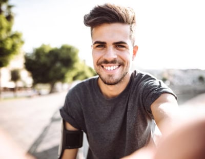 A young man with short, styled hair and a beard is smiling at the camera. He is wearing a gray T-shirt and an armband, likely for holding a phone or music player, while outside in a sunlit, tree-lined area. His arm is extended, suggesting he is taking a selfie.