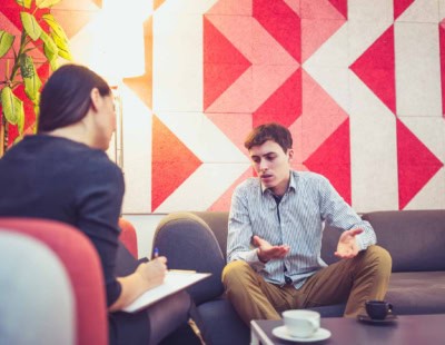 A man sits on a gray couch, gesturing with his hands while speaking to a woman who is taking notes on a clipboard. They are in a modern room with geometric red and gray wall patterns and a plant in the background. A coffee cup is on the table in front of them.