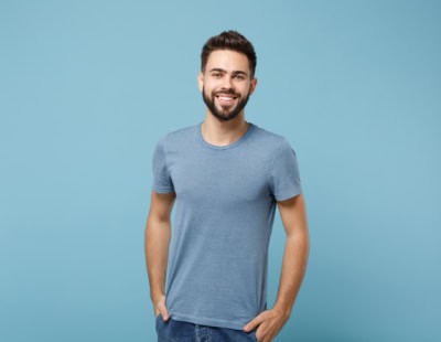 A man with a short beard and mustache, wearing a blue t-shirt and jeans, smiles while holding his hands in his pockets. He stands against a solid light blue background, embodying the approachable nature of outpatient treatment for professionals.