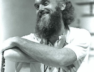 A black and white photo of a smiling person with a beard. They are leaning their arms on a raised knee and holding prayer beads, embodying the principles of Acceptance and Commitment Therapy. The person is wearing a white shirt and has a peaceful expression, with their hair pulled back. The background is out of focus.