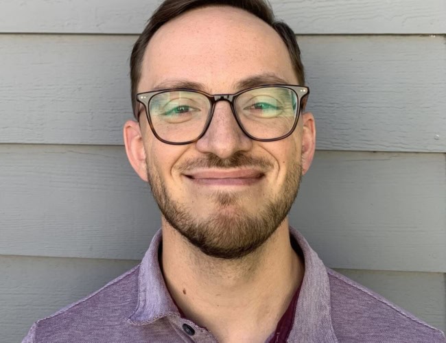 James Dowd, with short hair and glasses, smiles at the camera. He is wearing a purple collared shirt and standing in front of a gray wooden wall.