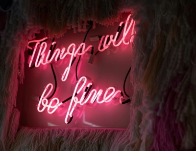 A neon sign with pink lighting displaying the message "Things will be fine," set against a textured, dark backdrop with soft, irregular fringes.