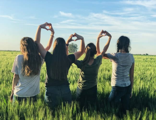 Five people stand in a grassy field facing away, forming heart shapes with their hands against a blue sky.